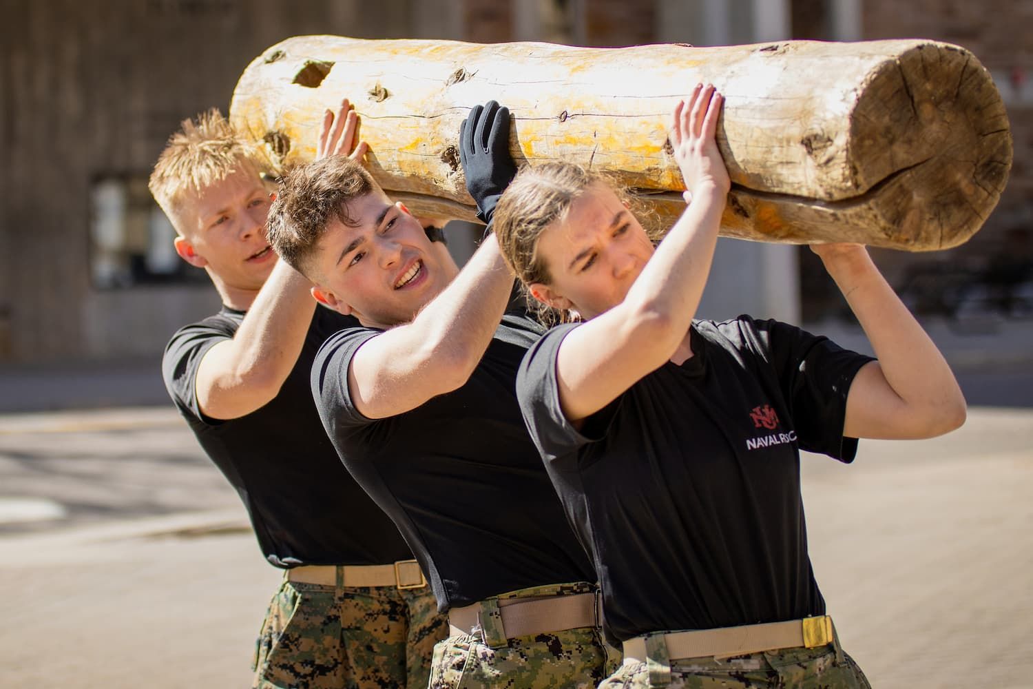 Three people lift a heavy wooden log outdoors; all wear black shirts and military-style pants, with strained expressions.