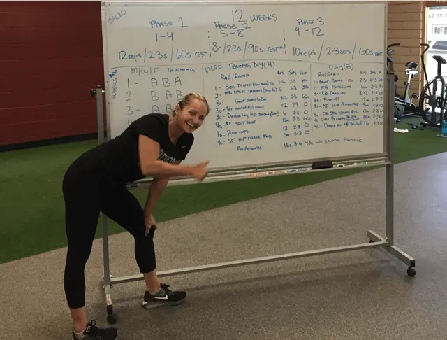 Woman giving a thumbs up next to a whiteboard with workout instructions in a gym.
