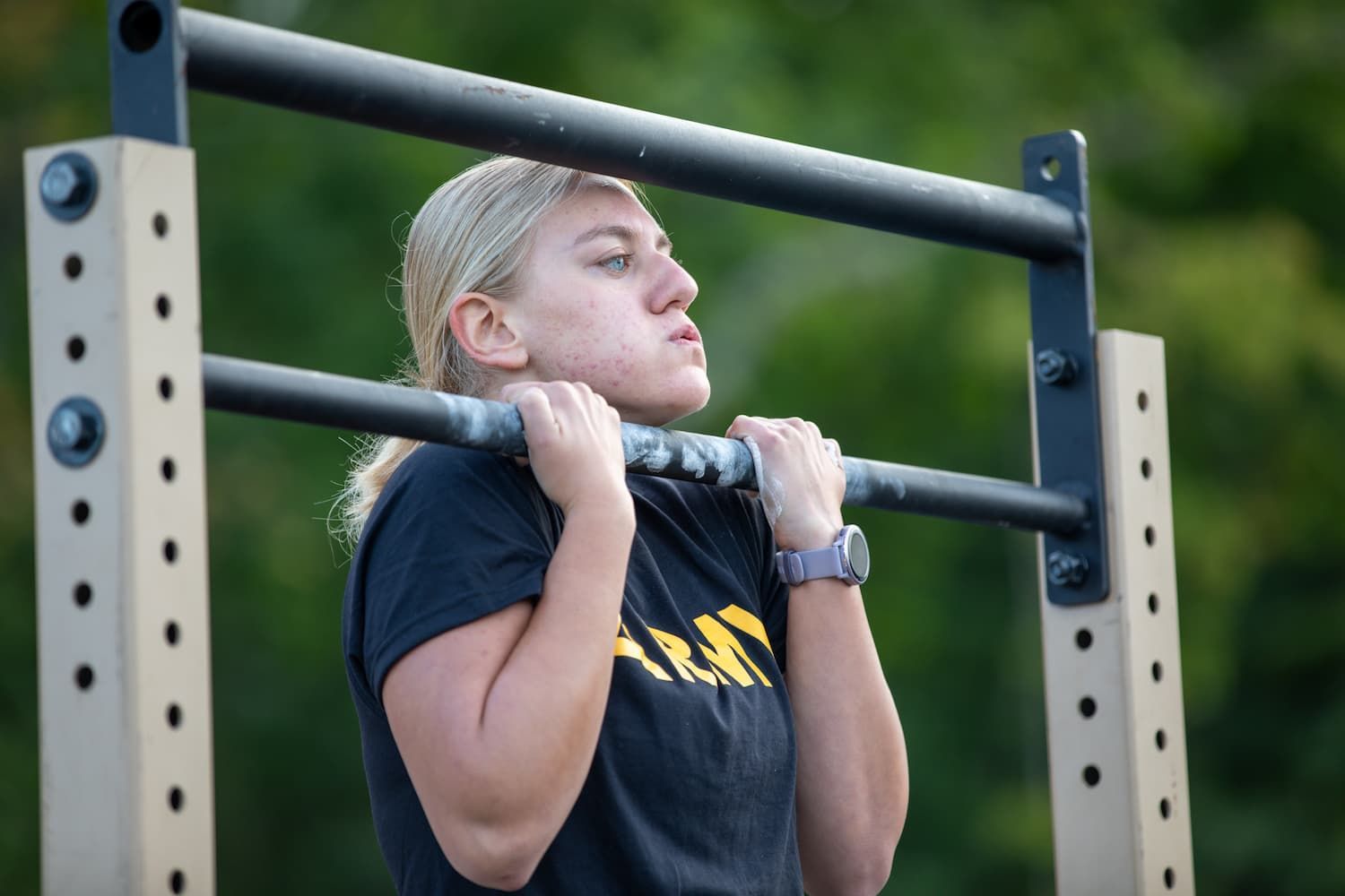 Woman in black shirt doing a pull-up on a metal bar outdoors, focused expression.