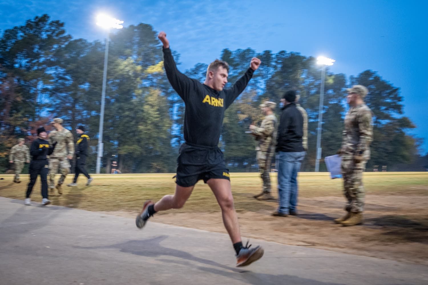 Soldier in black Army shirt and shorts raises arms, running, celebrating. Outdoor setting, other soldiers watching.
