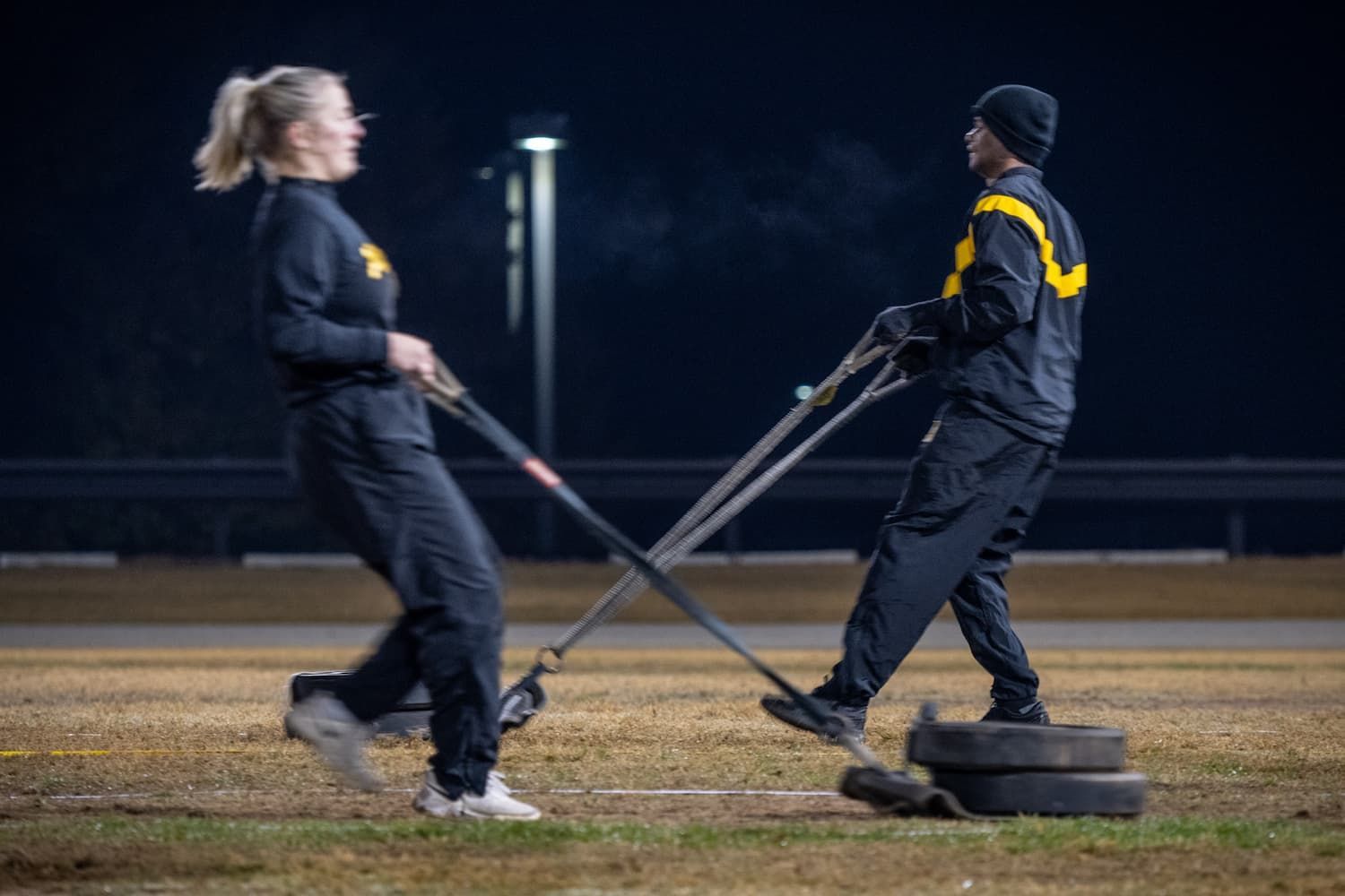 Two people, clad in black, pull a weighted sled on grass at night.