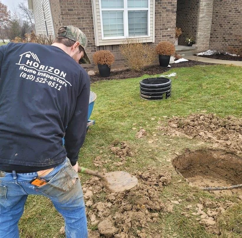 Worker installing a black drainage pipe beside a house, with a dug hole and dirt in the lawn.