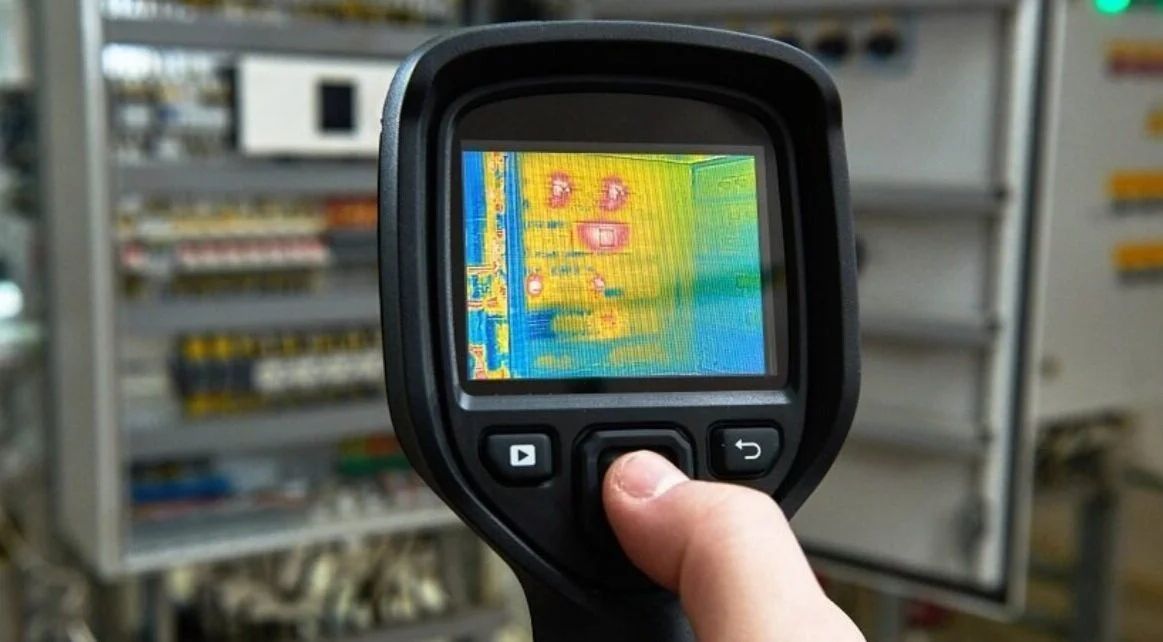 Handheld thermal camera scanning an electrical control panel, showing heat map colors on screen