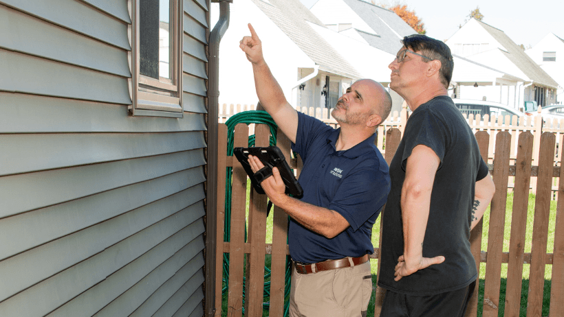 Two men inspect a house exterior; one points upward while the other holds a tablet near the siding.
