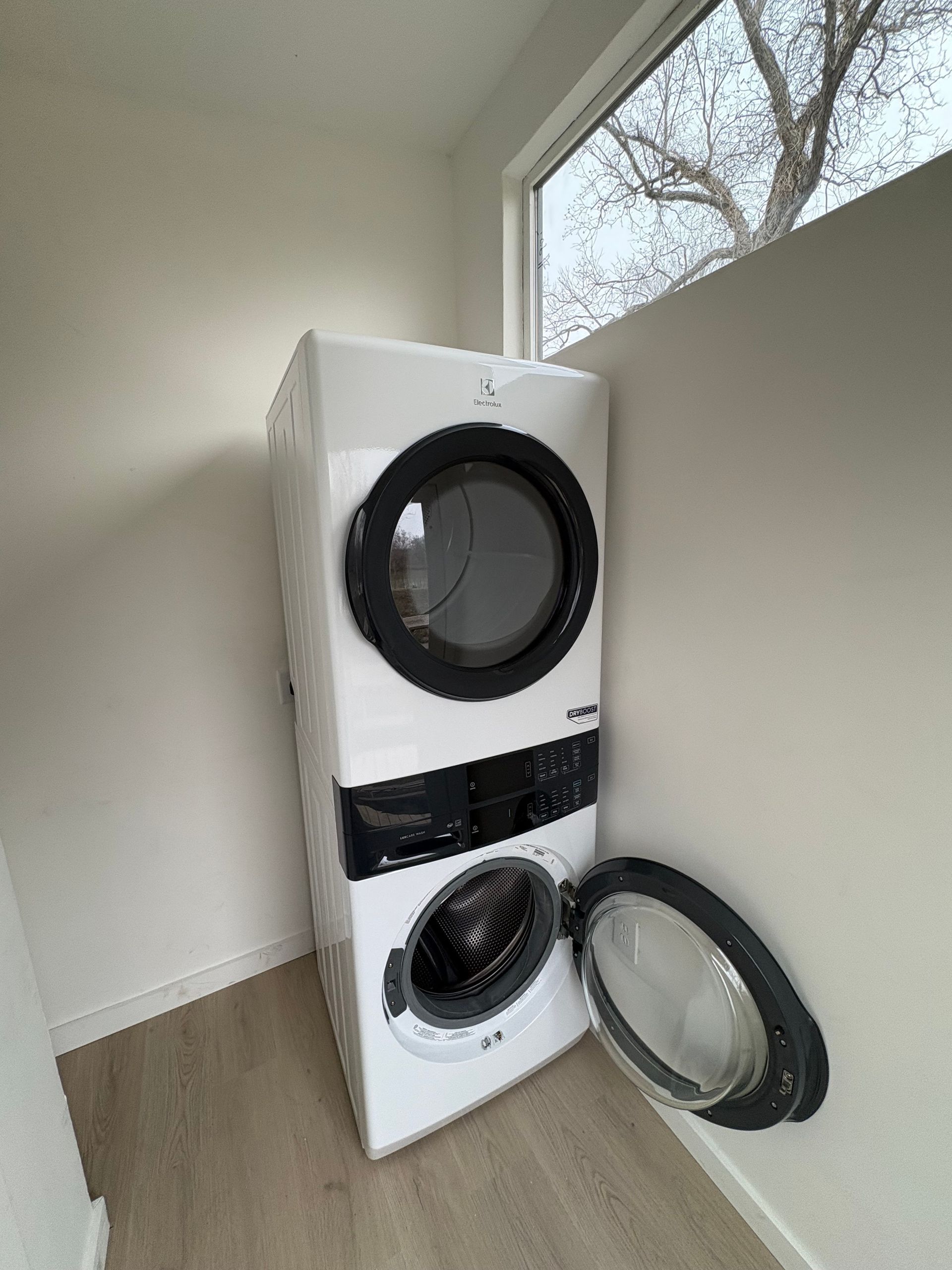 Stacked washer and dryer in a bright room, with both appliance doors open and leaning against a wall.