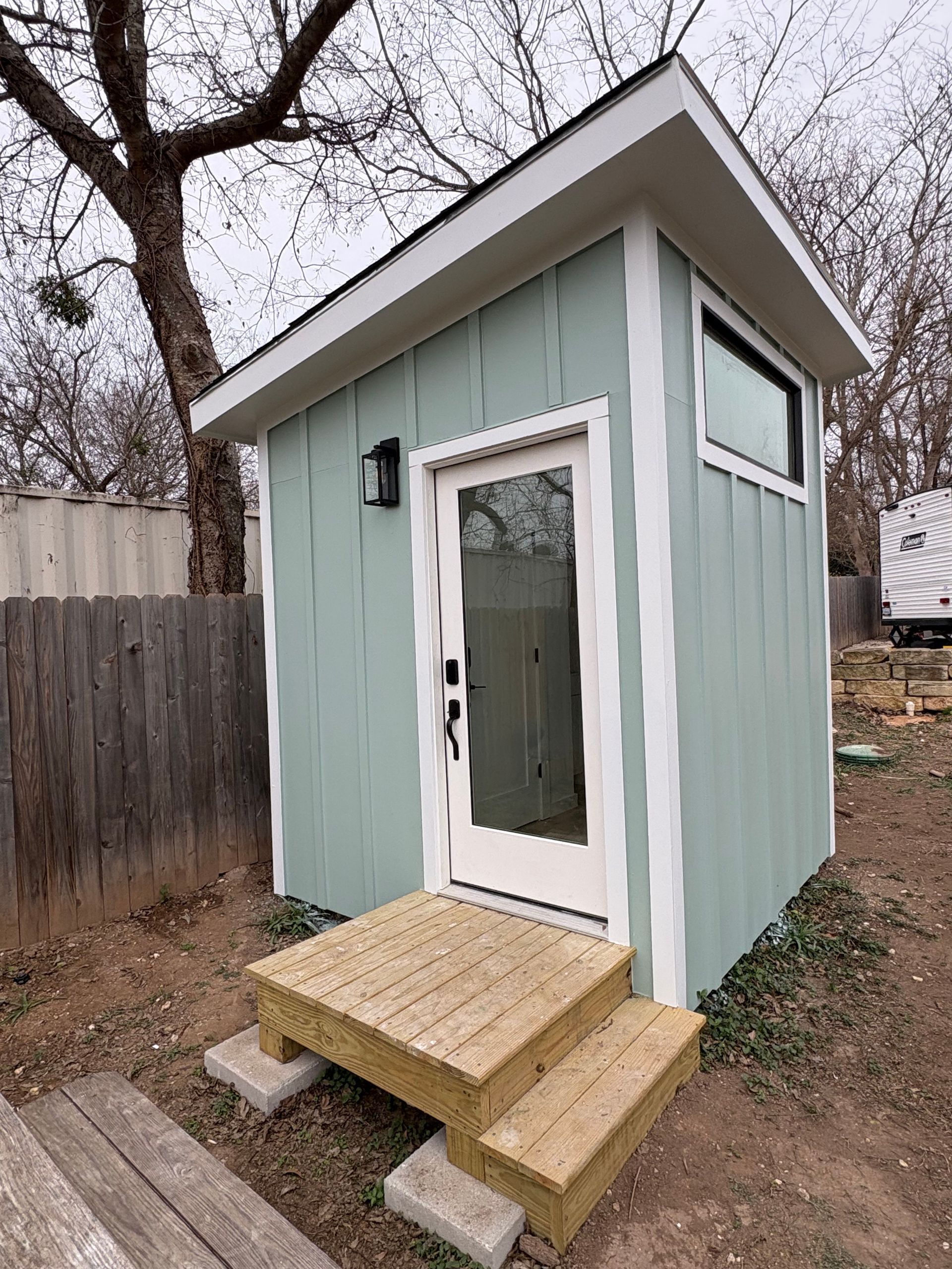 Small mint-green backyard shed with white door and steps beside a wooden fence.