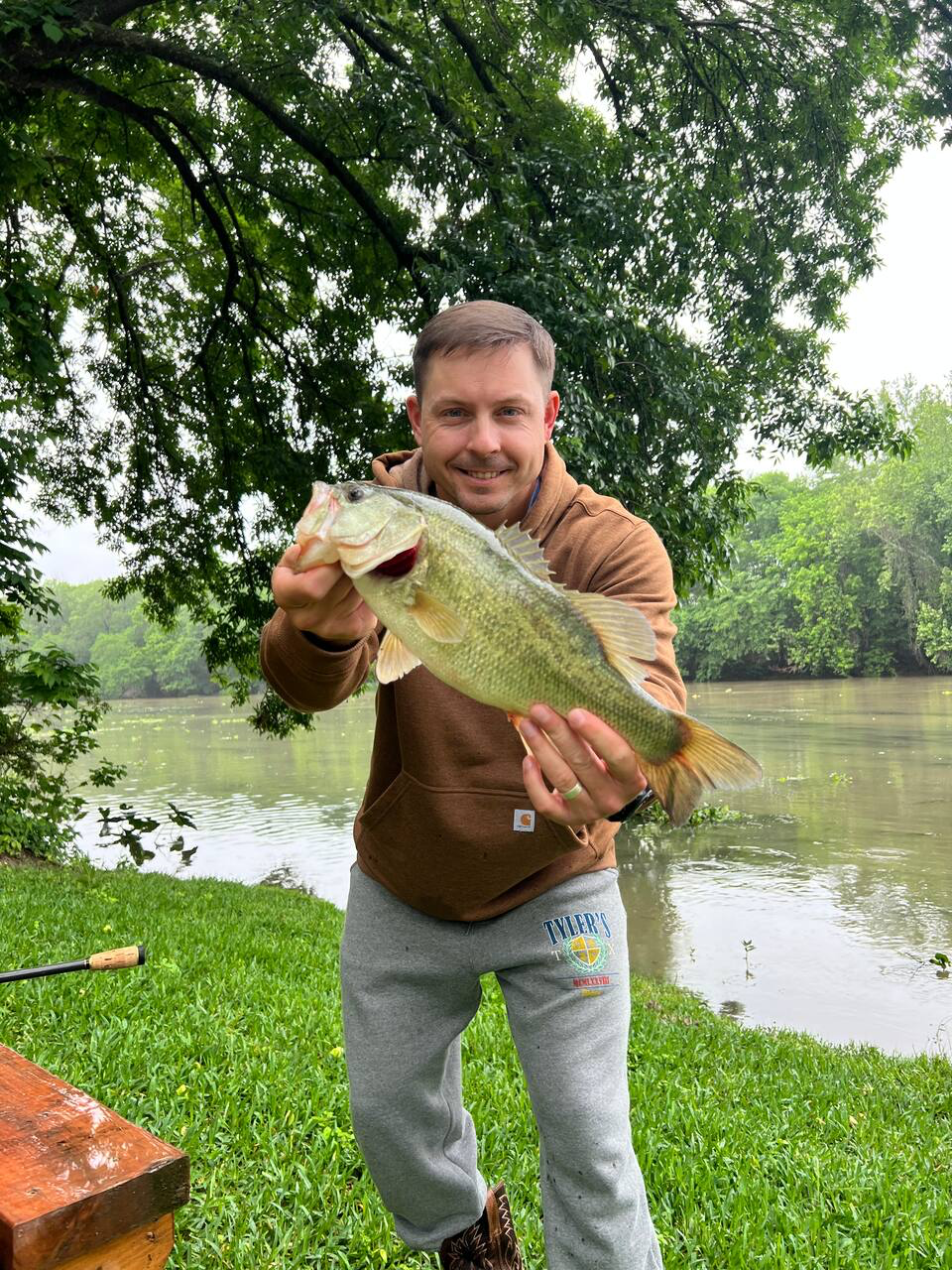 Man holding a large fish by a river, standing on bright green grass under a leafy tree.