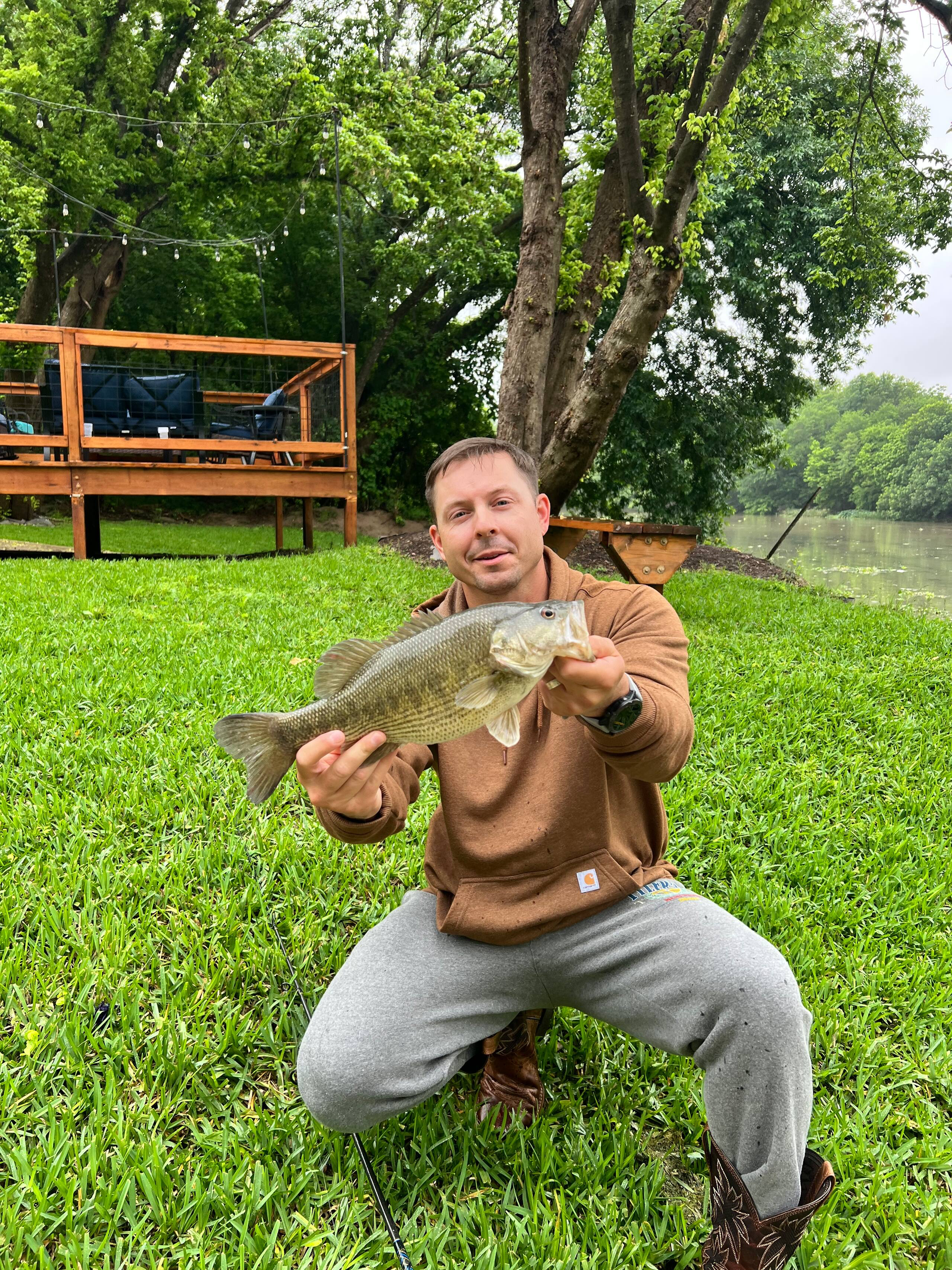 Man kneeling by a pond holding a large fish, with a wooden deck and lush greenery behind him.
