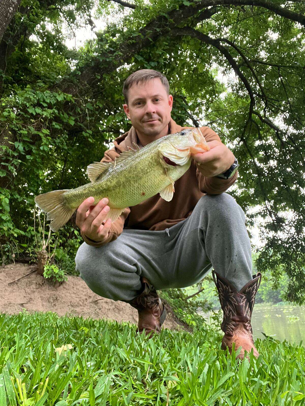 Man crouching outdoors, holding a large fish near a grassy bank and trees.