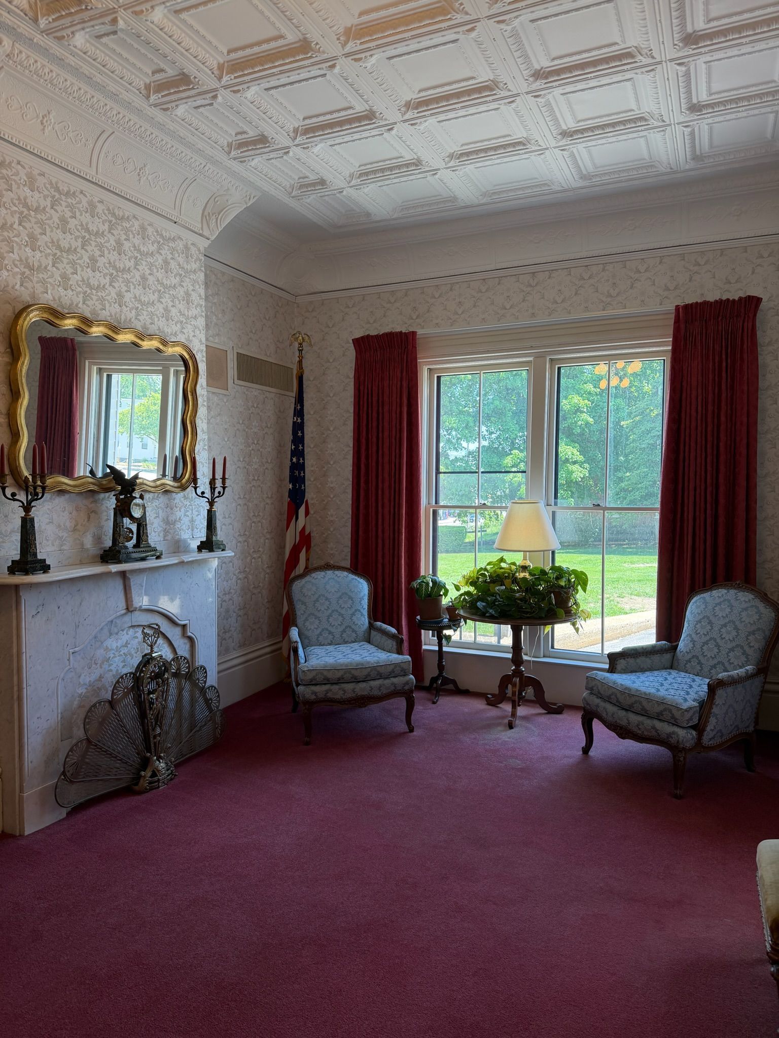 Elegant sitting room with red carpet, ornate white ceiling, fireplace, and large windows overlooking a garden