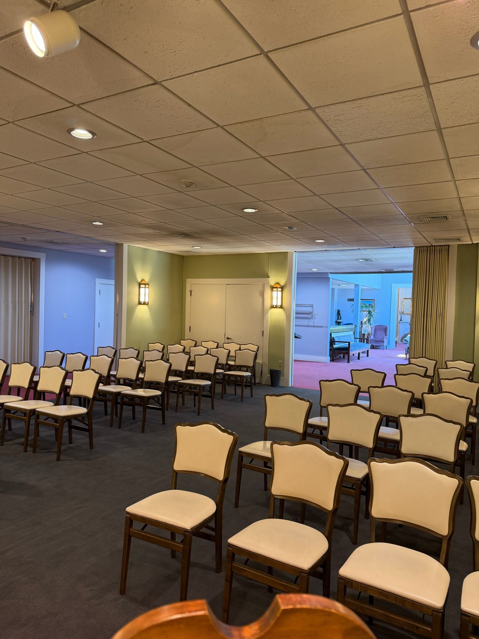 Empty conference room with rows of beige chairs, carpeted floor, and ceiling lights, viewed toward a bright doorway.