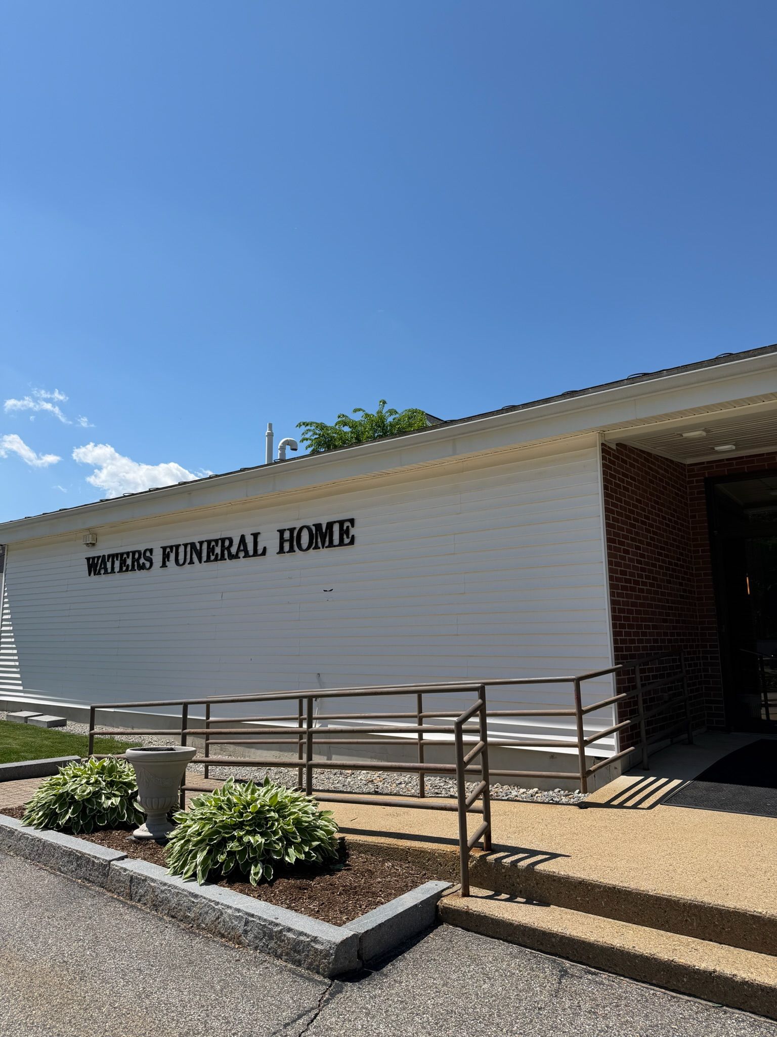 White funeral home building with ramp entrance and landscaped flower bed under a clear blue sky