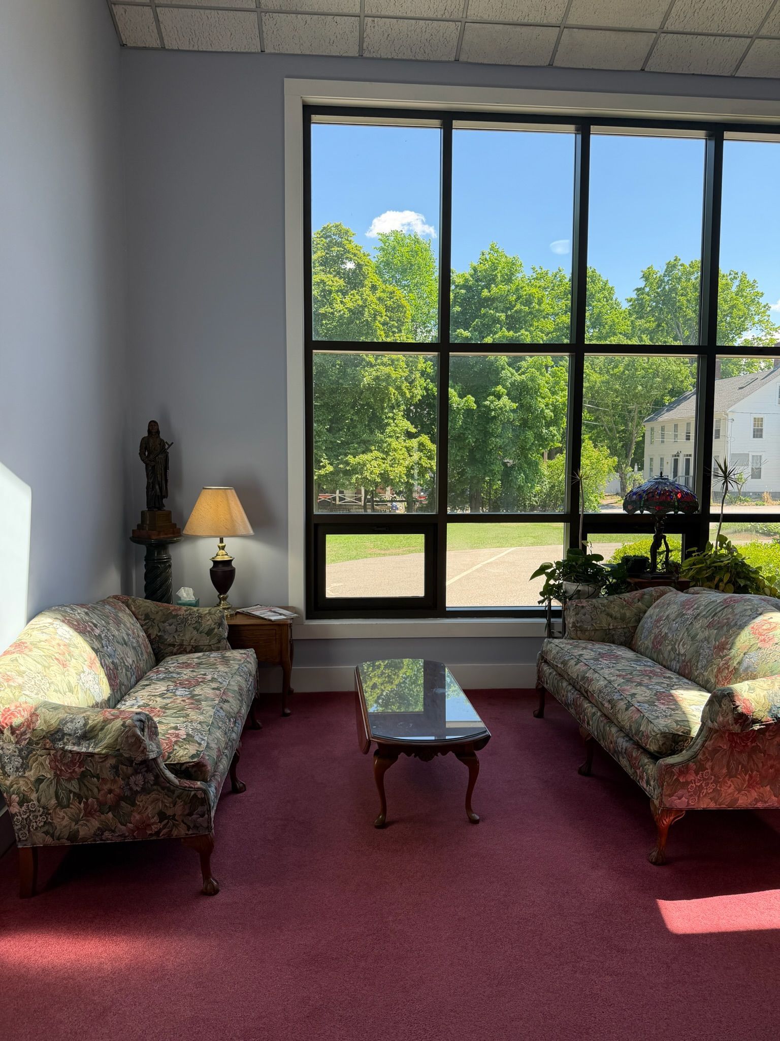 Sunlit living room with two floral sofas, red carpet, coffee table, lamp, and large window overlooking trees