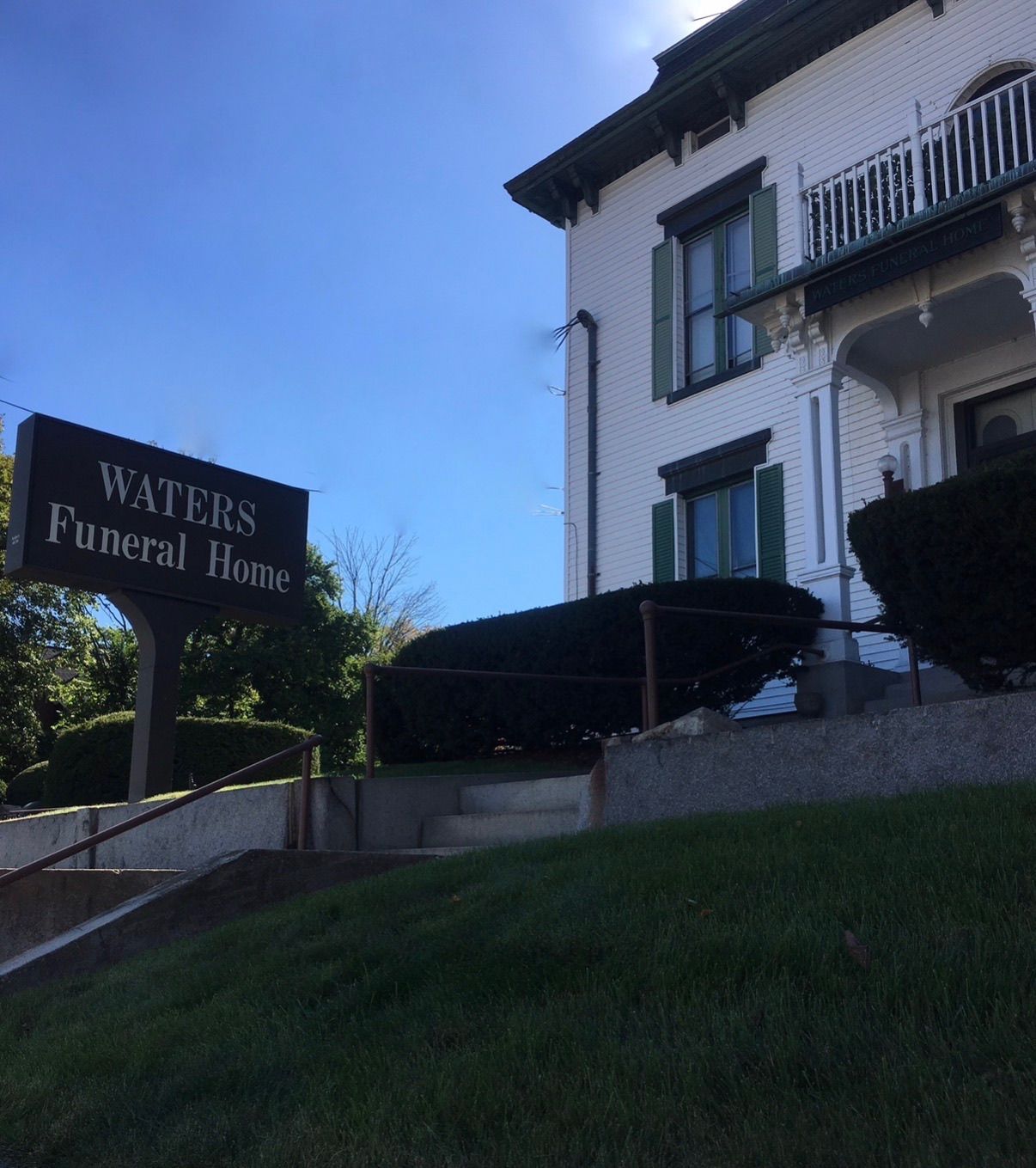 Waters Funeral Home sign beside a white building with green shutters on a grassy slope