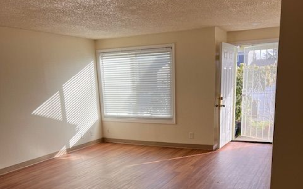 Empty room with closed blinds, open door, sunlight, and wood-look flooring.