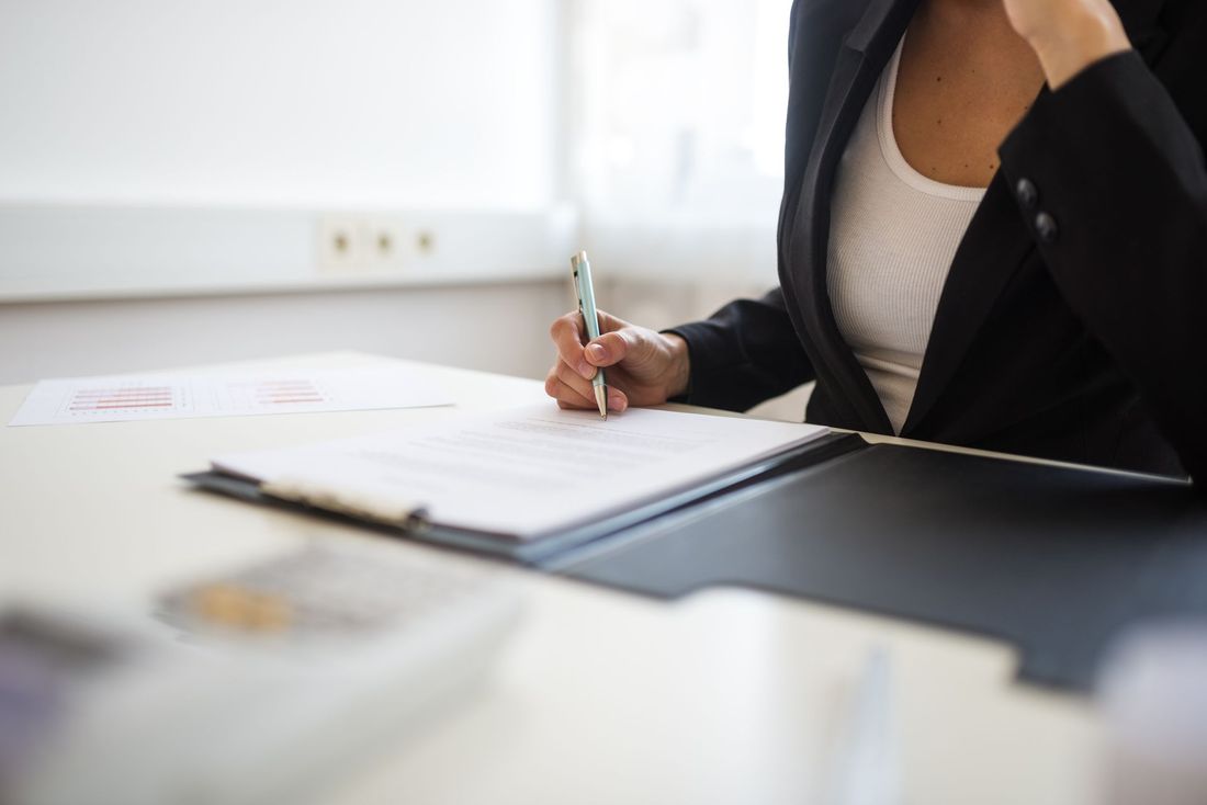 Una persona con un blazer nero firma dei documenti a una scrivania in un ufficio luminoso.