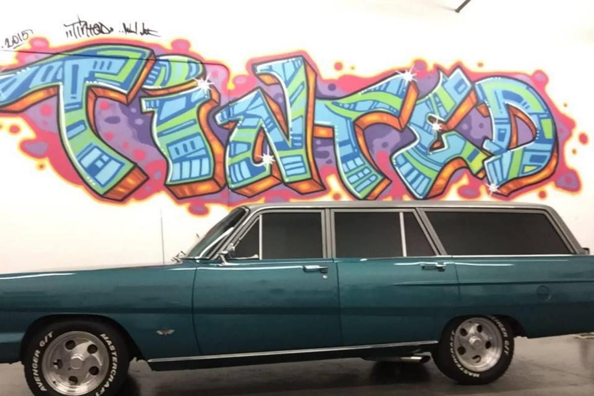 A blue car is parked in front of a wall with graffiti that says trusted