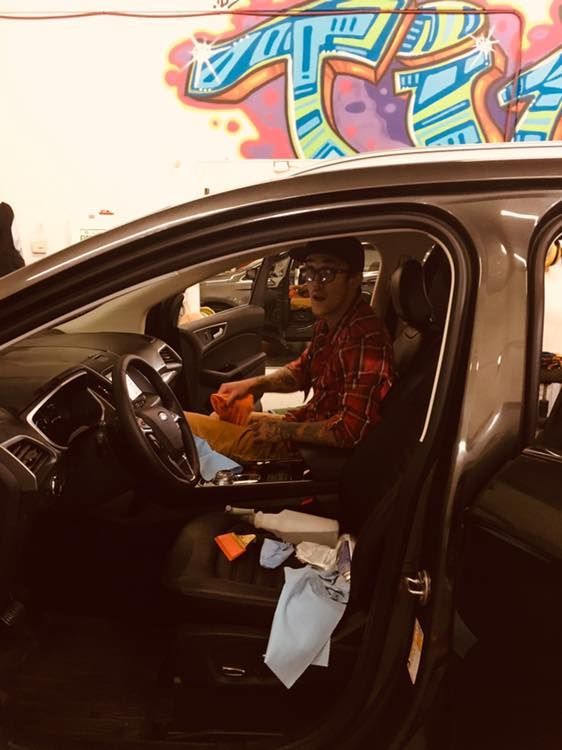 A man is sitting in a car with graffiti on the wall behind him.
