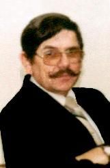 Portrait of a man in a dark suit, glasses, and a mustache, posing with folded arms against a plain background