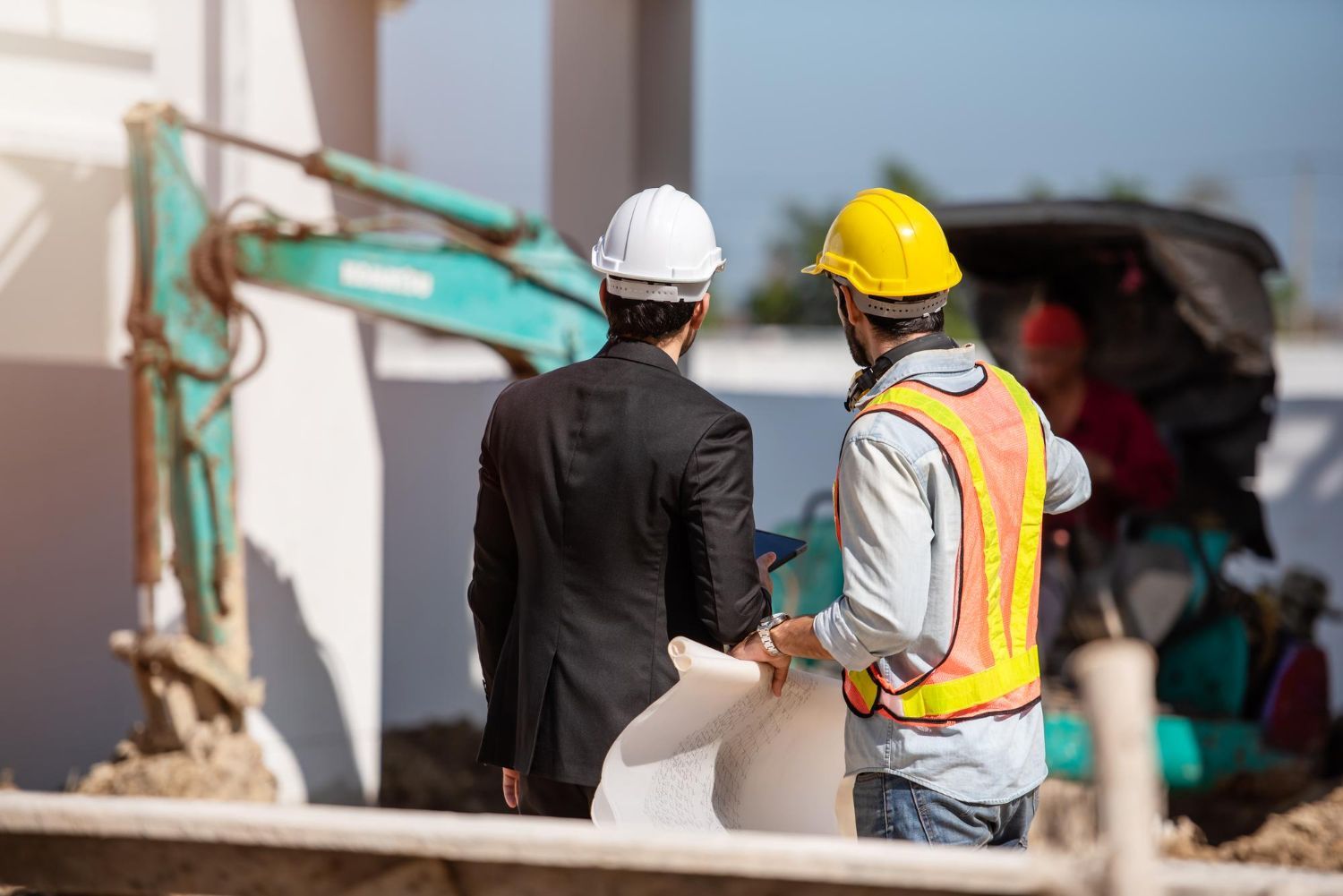 Two men in hard hats examine blueprints at a construction site near a small excavator.