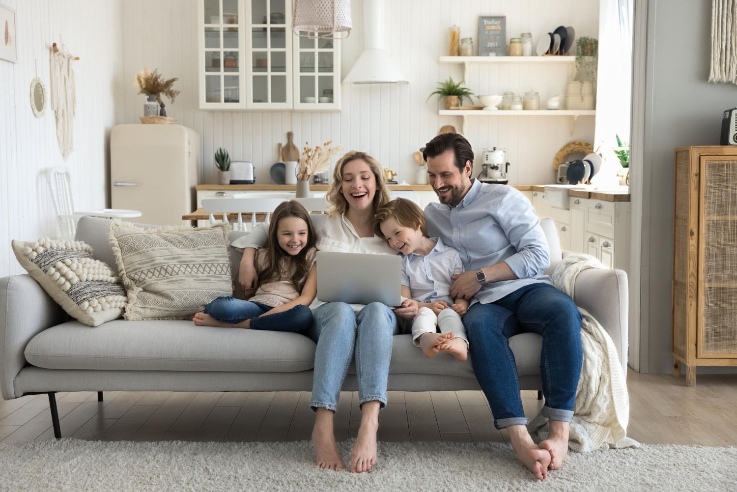 Family of four laughs while watching a laptop on a living room couch.