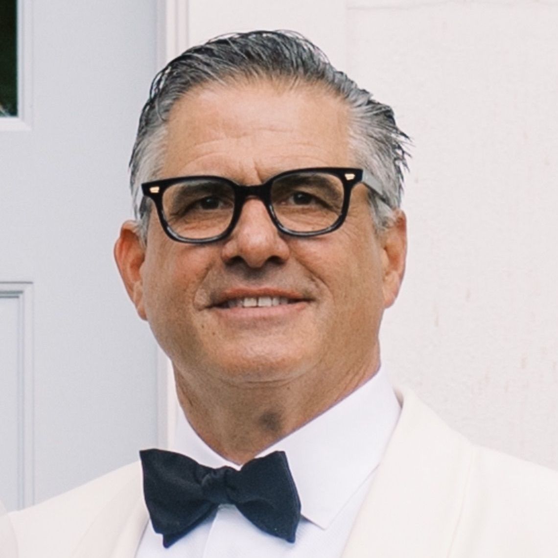 Man in black glasses and bow tie, smiling, in front of a white building.