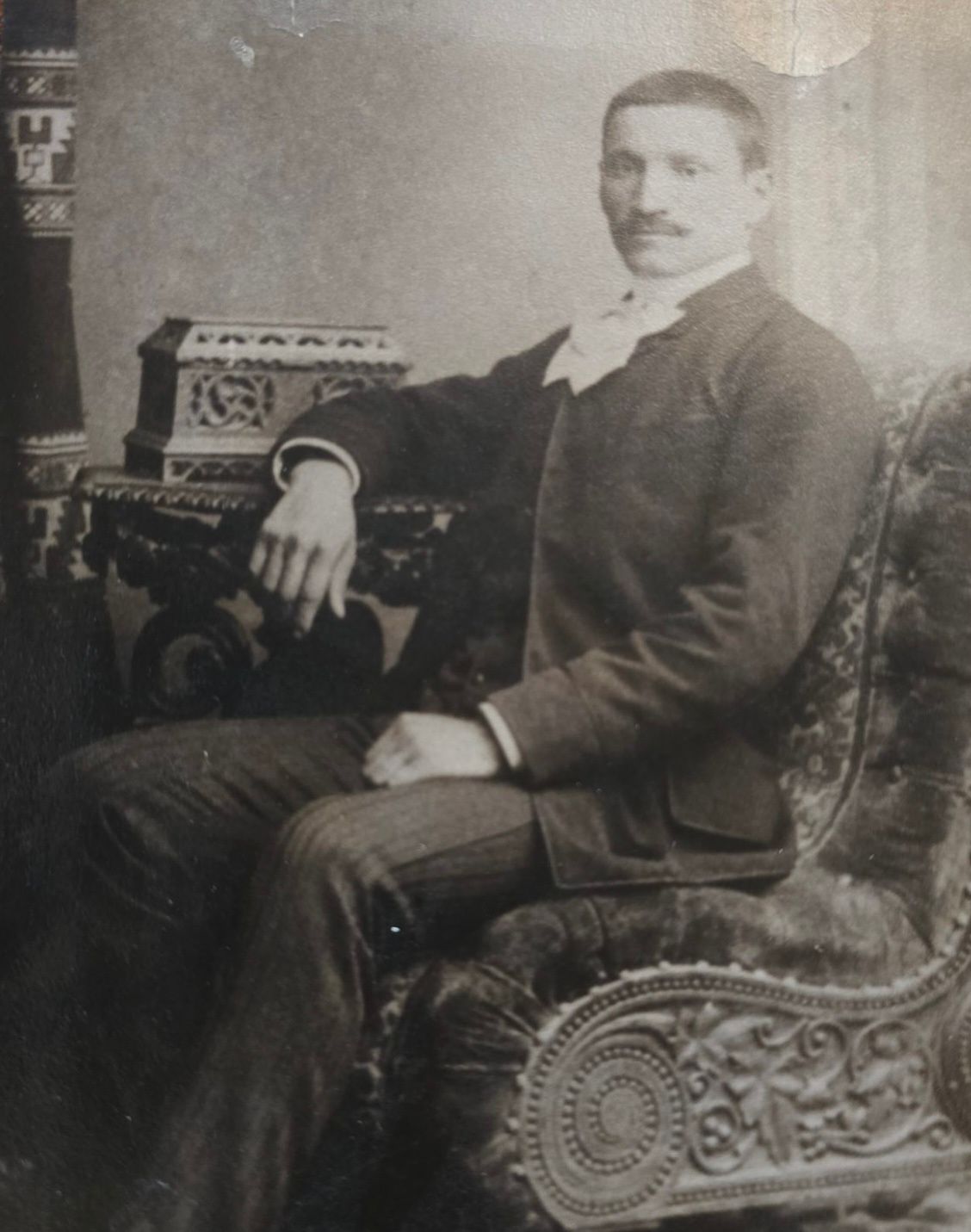 Man in suit seated on ornate chair, resting arm on decorated table.