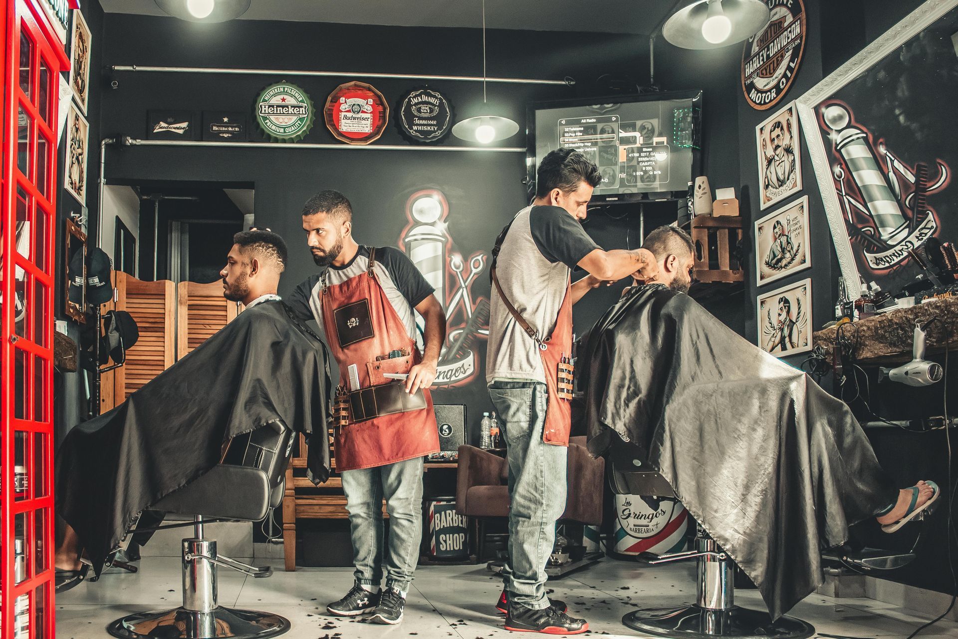 Two barbers giving haircuts in a vintage-style barbershop with dark walls.