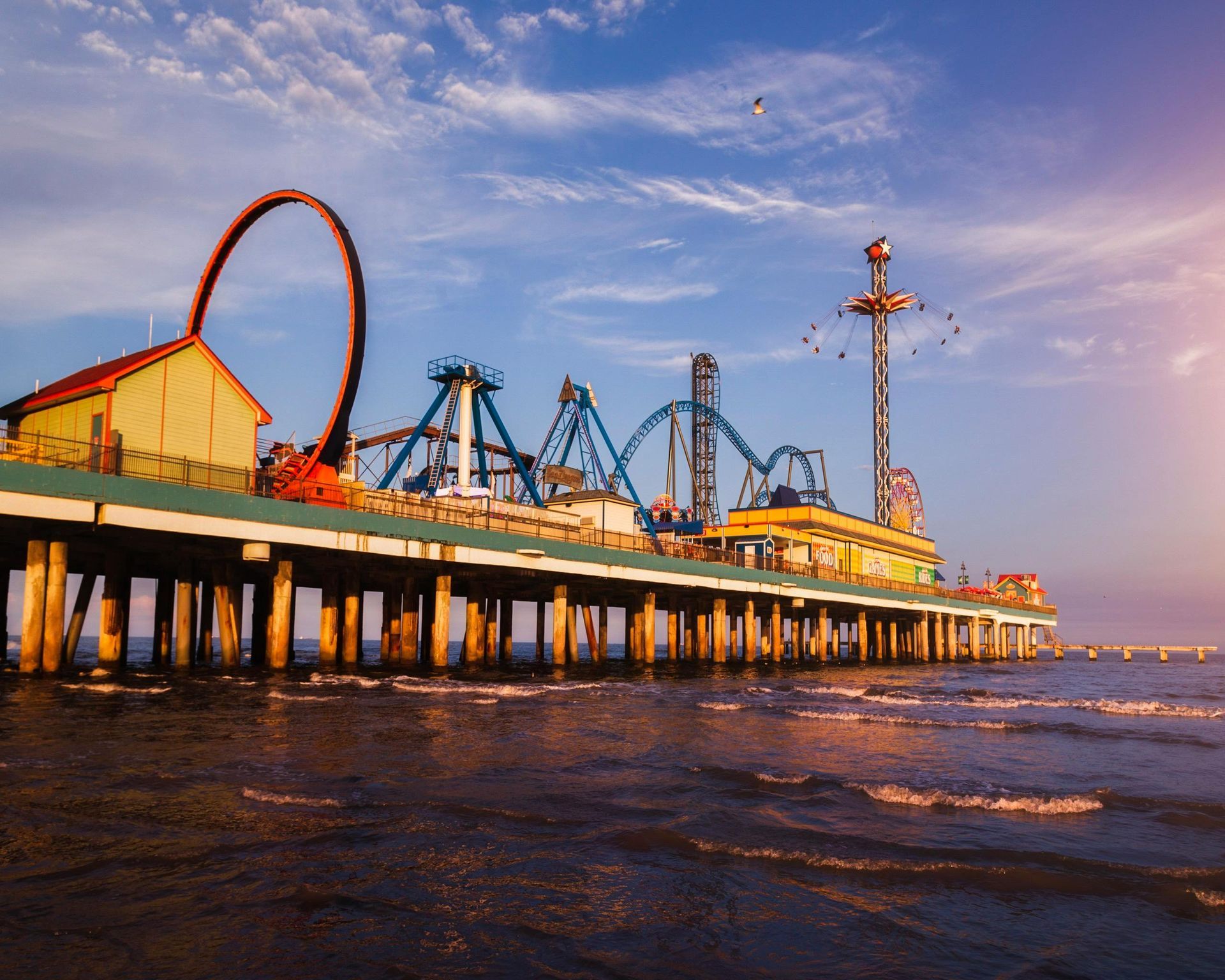 Pleasure pier amusement park with roller coasters and rides at sunset, extending over the ocean waters.