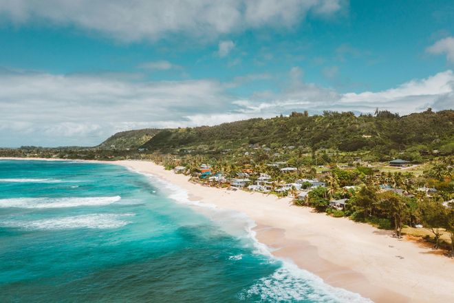 Beach with turquoise water, white sand, and lush green hillside under a blue sky with clouds.