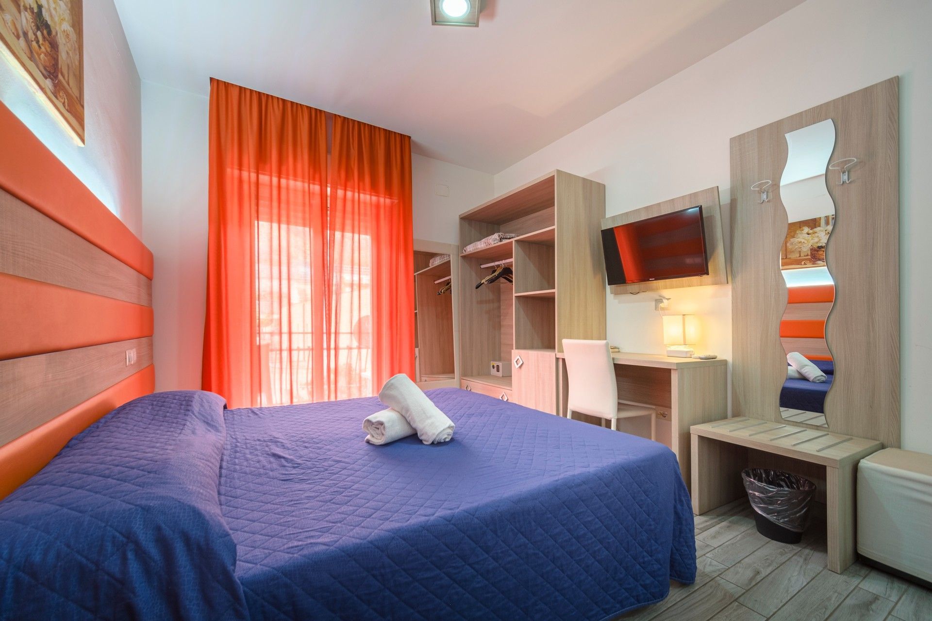 Una camera d'albergo con un letto viola, tende arancioni, una scrivania e un televisore.