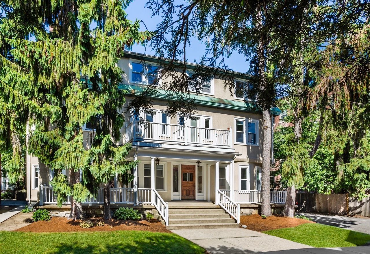 Two-story beige apartment building with a front porch, stairs, and tall trees surrounding.