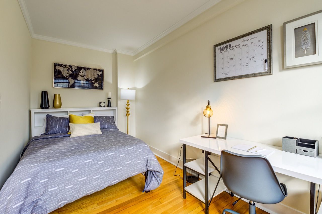Bedroom in an apartment featuring a bed, white desk, chair, and decorative wall calendar.