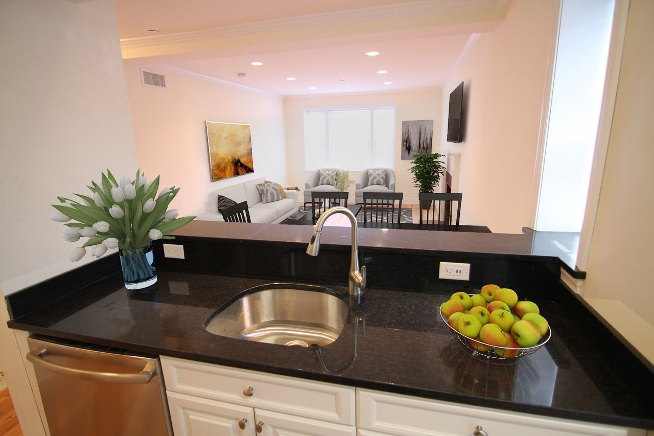 Open-concept kitchen with a black granite countertop, stainless sink, and a fruit bowl, facing a bright living area.
