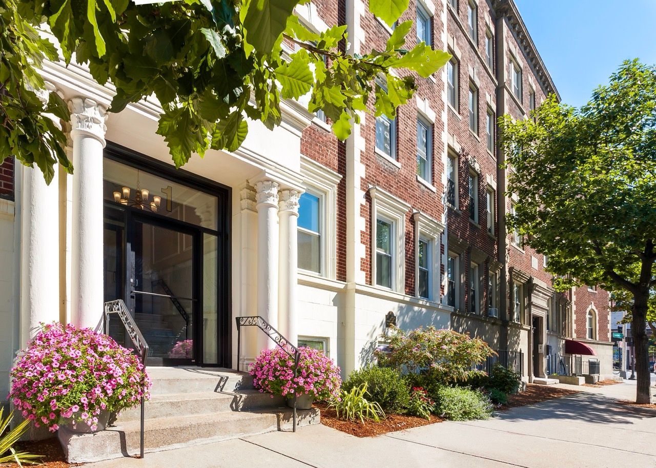 Brick apartment building entrance with white columns, steps, and flowering planters.