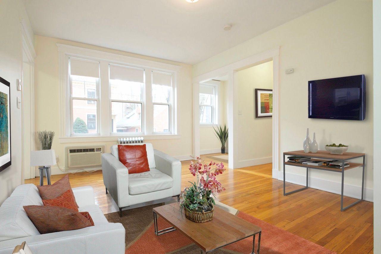 Bright apartment living room with white sofas, orange cushions, wood floors, large windows, and TV.