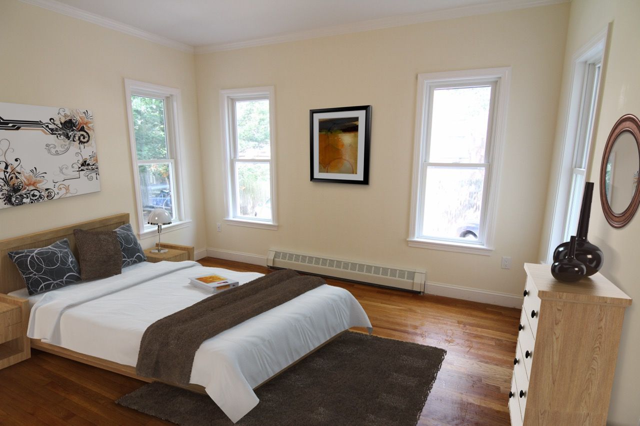 Bright bedroom with wooden bed, dresser, round mirror, and three windows.