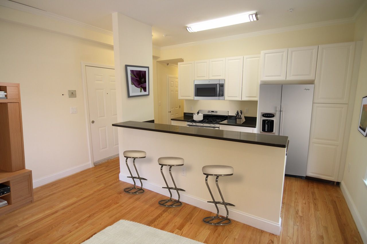 Open kitchen with white cabinets, stainless steel refrigerator, microwave, and a breakfast bar with three white stools.