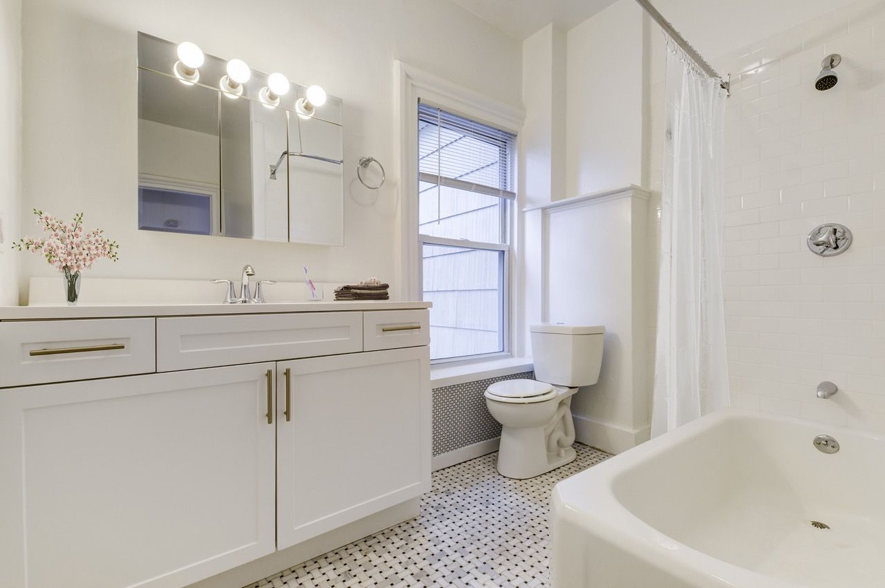 Bright white apartment bathroom with vanity, large mirror, toilet, window, and shower/tub combo.