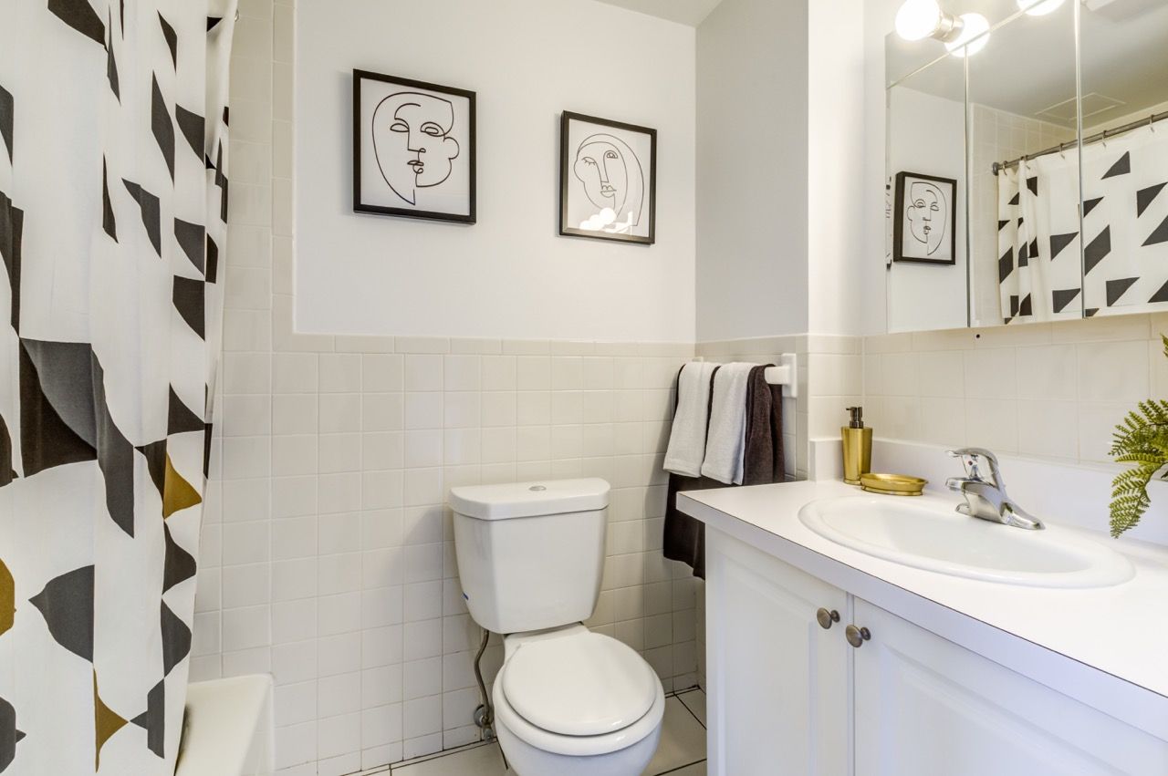 White-tiled apartment bathroom with toilet, sink, mirror, and geometric shower curtain.