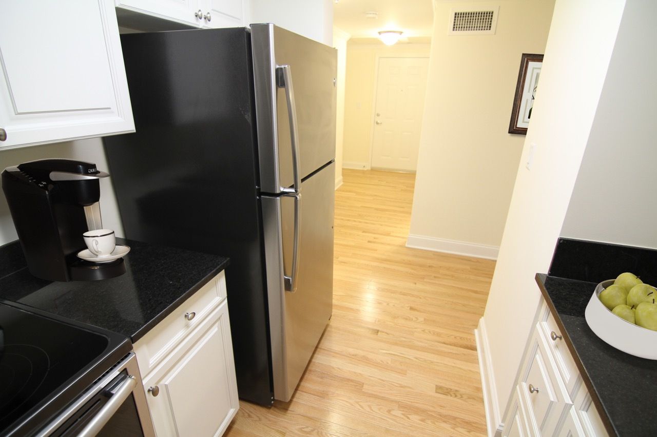 Kitchen in apartment with stainless steel refrigerator, black countertops, white cabinets, and light hardwood floors.