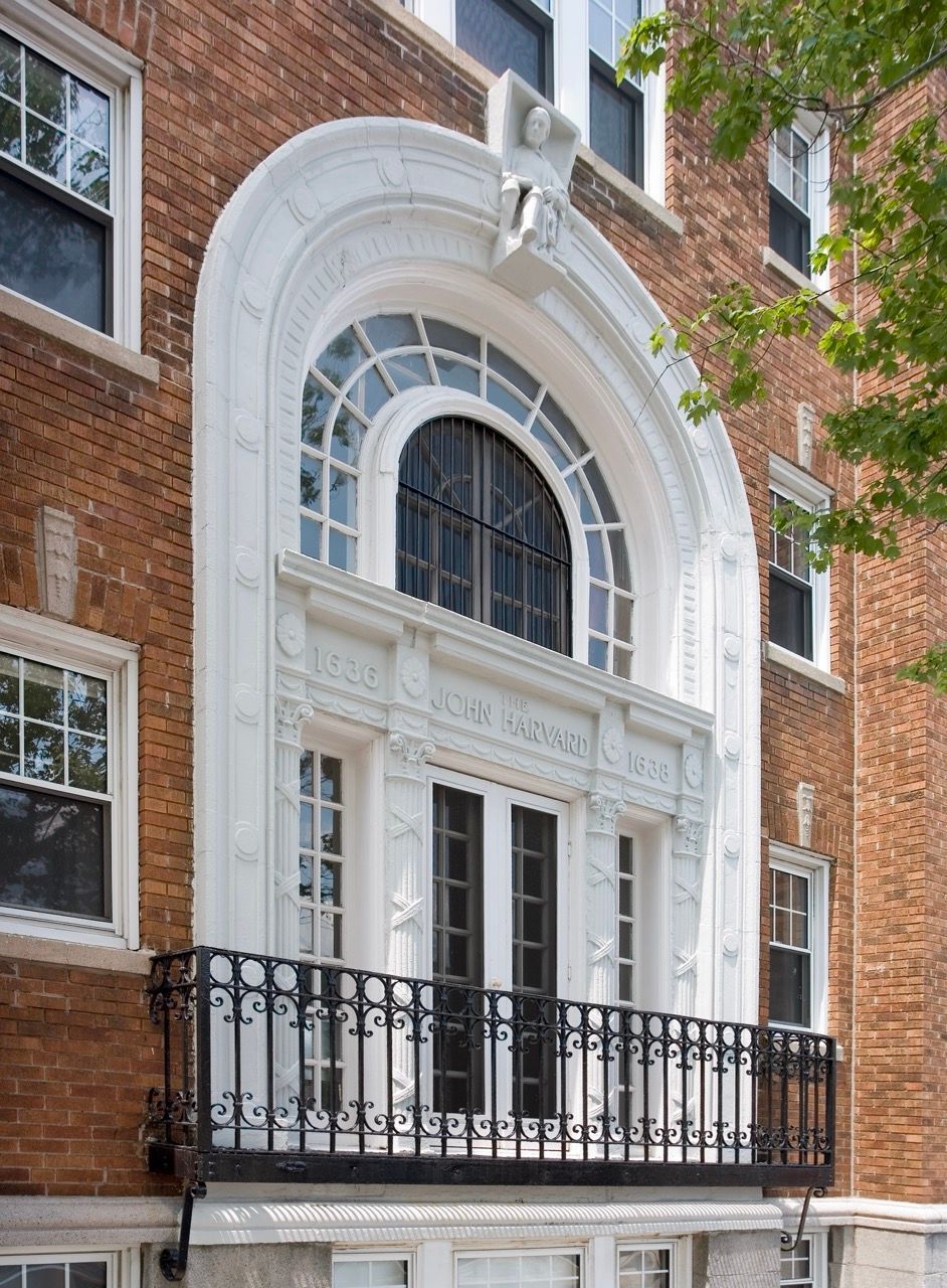 Brick apartment building exterior with a white ornate arched entry and black wrought-iron balcony.
