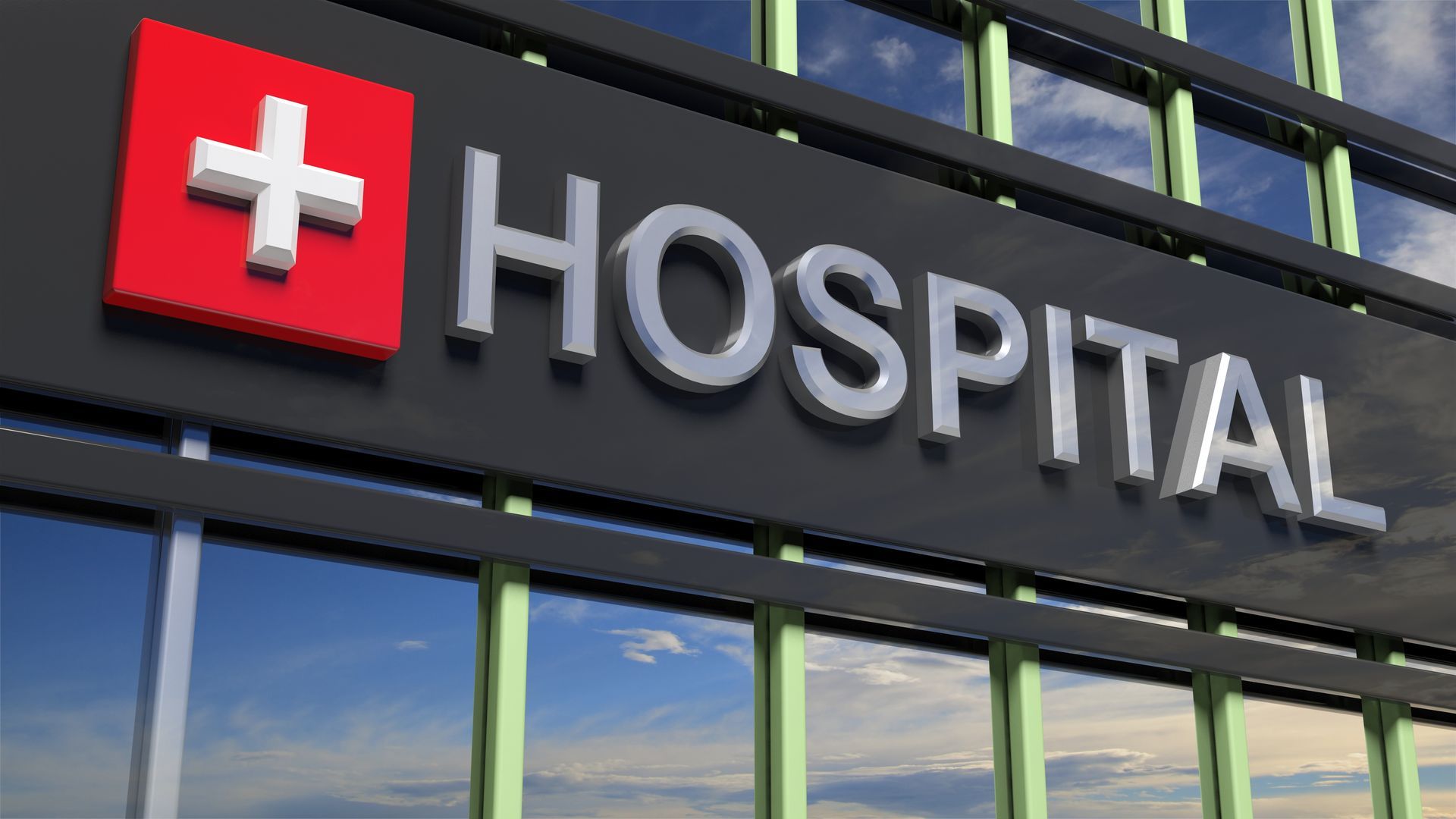 Hospital sign with a white cross on a red square and bold letters on a glass building.