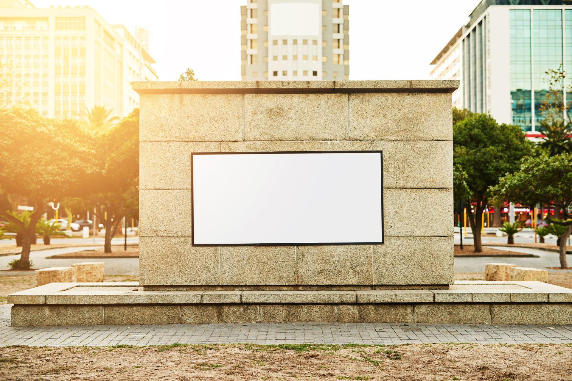 Blank billboard on a stone pedestal in a sunny city plaza with buildings in the background