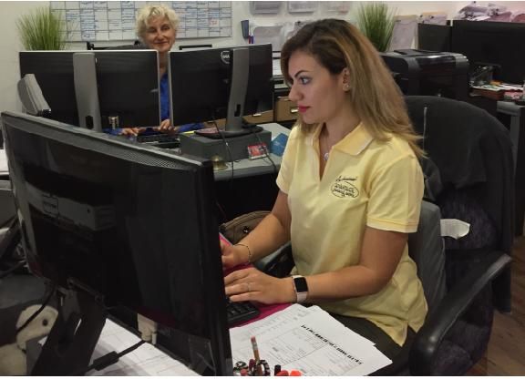 Woman in yellow shirt types at computer, another woman works in background. Office setting.