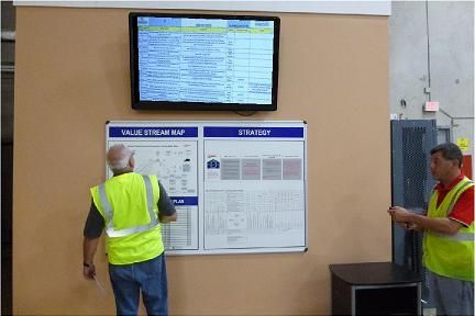 Two men in safety vests review charts on a wall in an industrial setting, with a monitor above.