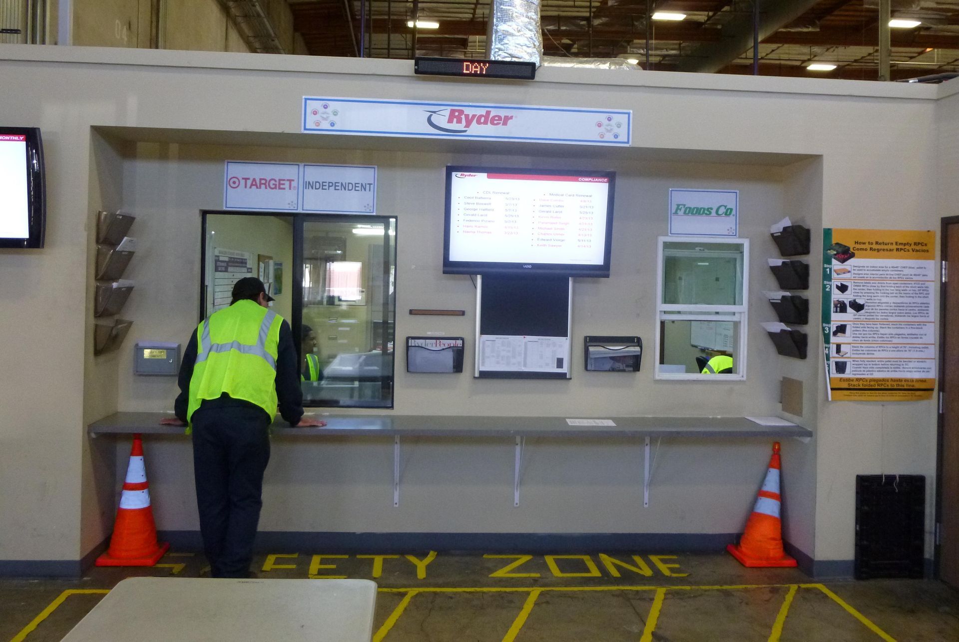 Worker in a yellow vest at a factory safety station. A screen displays information, with orange cones.
