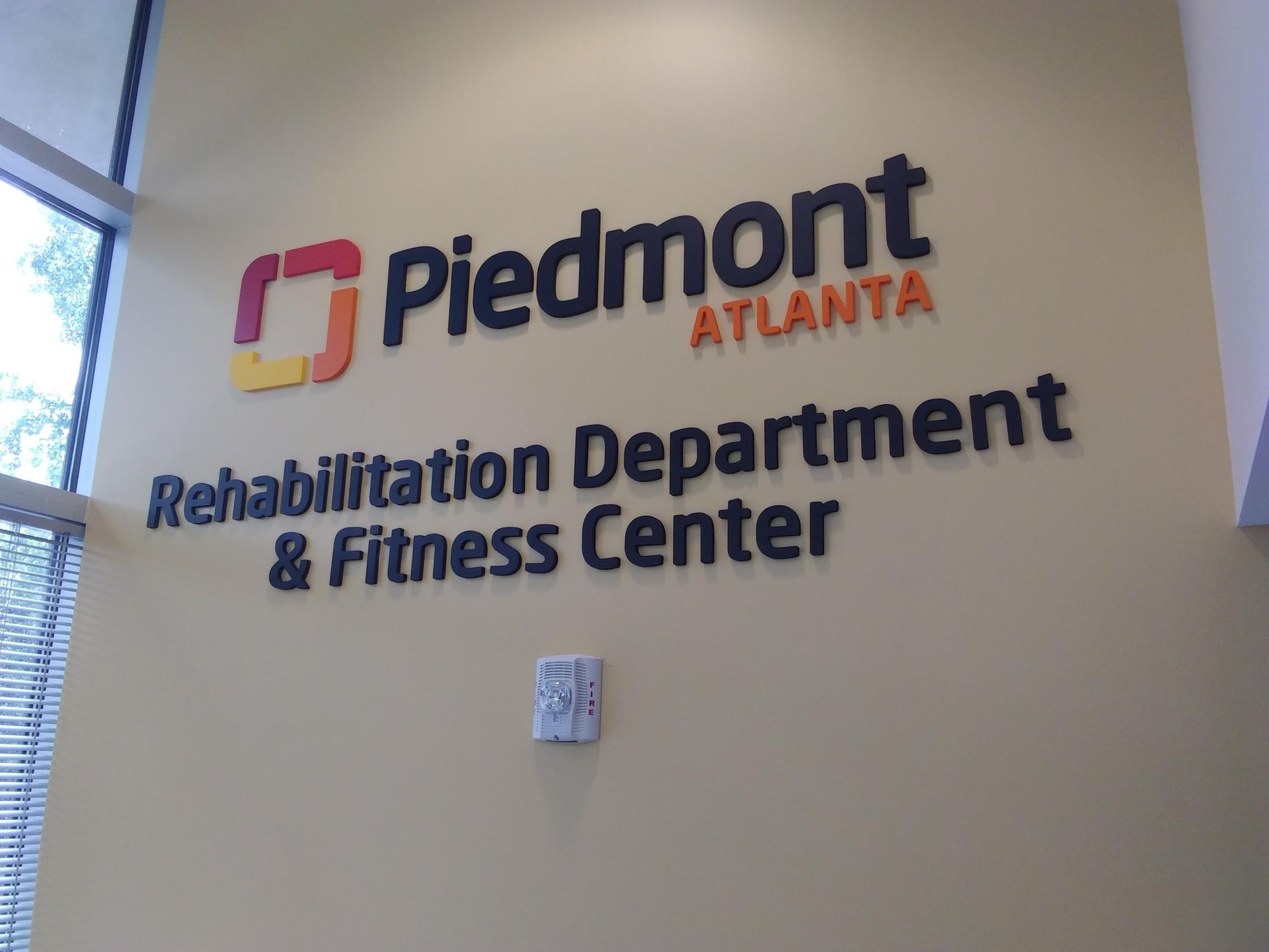 Sign for Piedmont Atlanta Rehabilitation Department & Fitness Center on a tan wall. The Piedmont logo is orange/red.