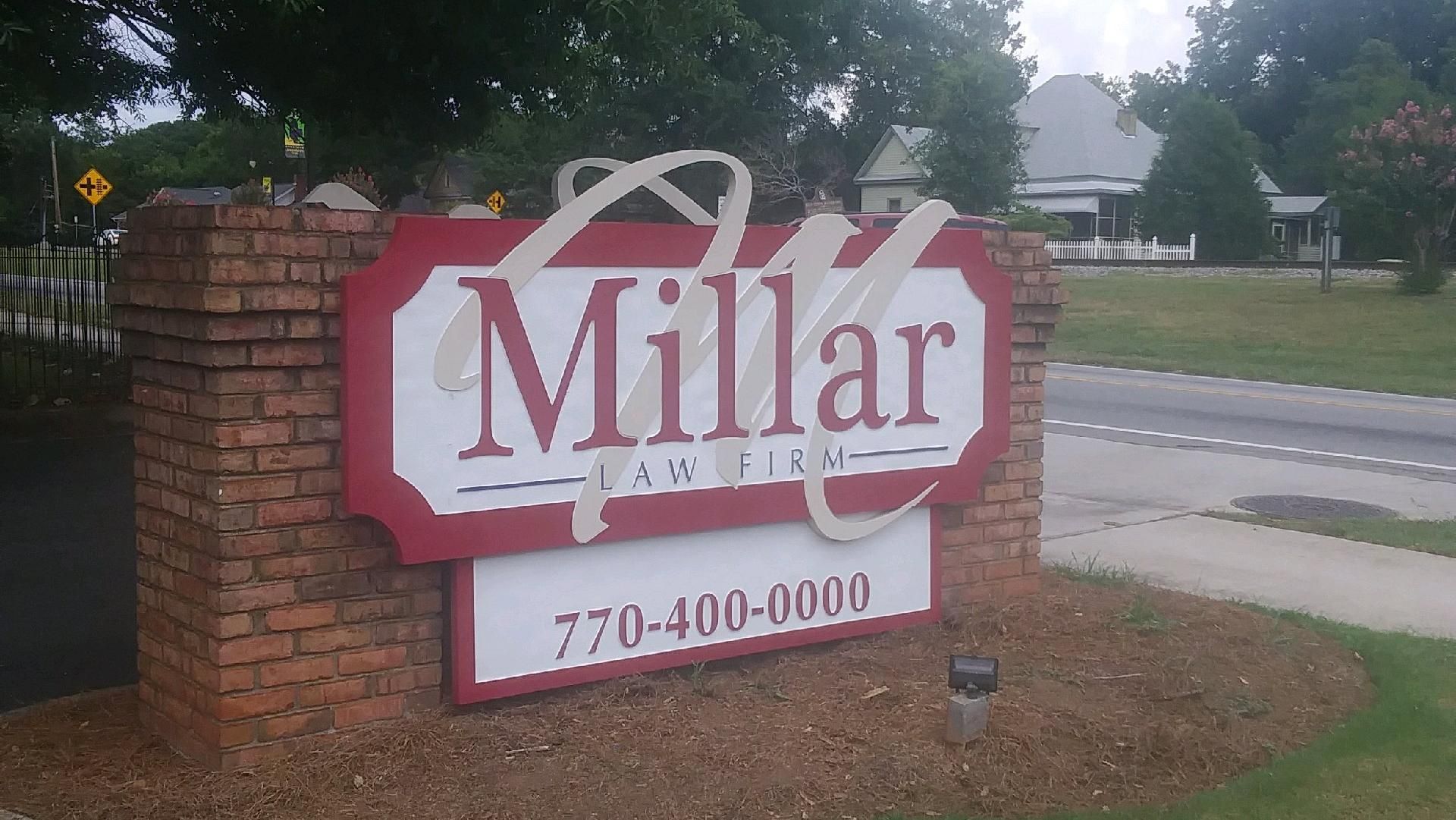 Sign for Millar Law Firm, brick structure, red and white lettering with a phone number and building in the background.