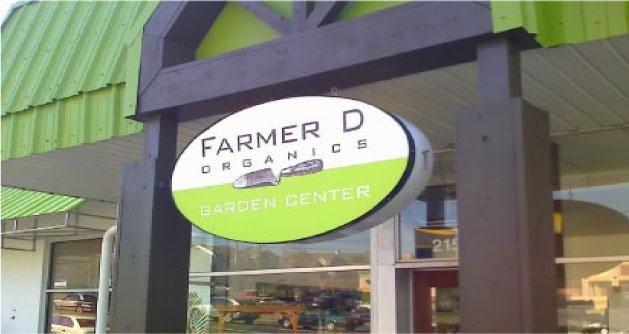 Sign for Farmer D Organics Garden Center: oval, white and green with brown accents, over a storefront.